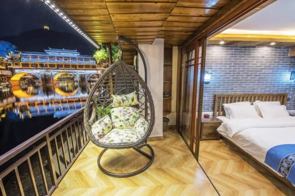 Quarto com uma cama, cadeira suspensa, piso de madeira e vista para um edifício tradicional.