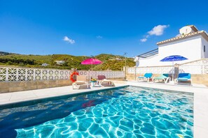 Une piscine à l’eau d’un bleu cristallin, entourée de murs blancs et d’une clôture blanche. On y trouve des chaises longues et des parasols, et une colline verdoyante en arrière-plan.