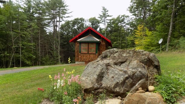 Una cabaña pequeña con techo rojo y detalles verdes, rodeada de un paisaje natural con rocas grandes y plantas con flores.