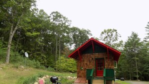 Una cabaña pequeña con techo rojo, fachada de madera y puerta verde. Se ve una parrilla y una silla afuera.