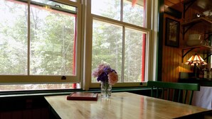 Una mesa de madera con un florero, un libro y una ventana que da a vista de árboles.