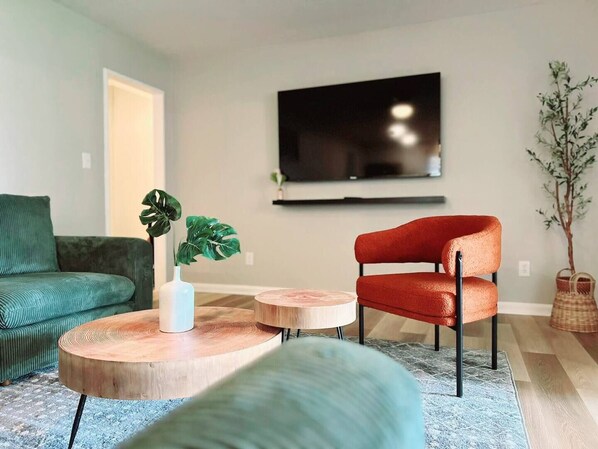 Un salon avec un canapé vert, un fauteuil rouge, une table basse et un téléviseur fixé au mur.