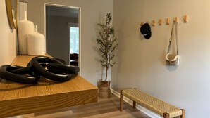 Een moderne kamer met een houten bankje, een klein tafeltje met zwarte, opgerolde objecten, een potplant en een kapstok met hoeden.