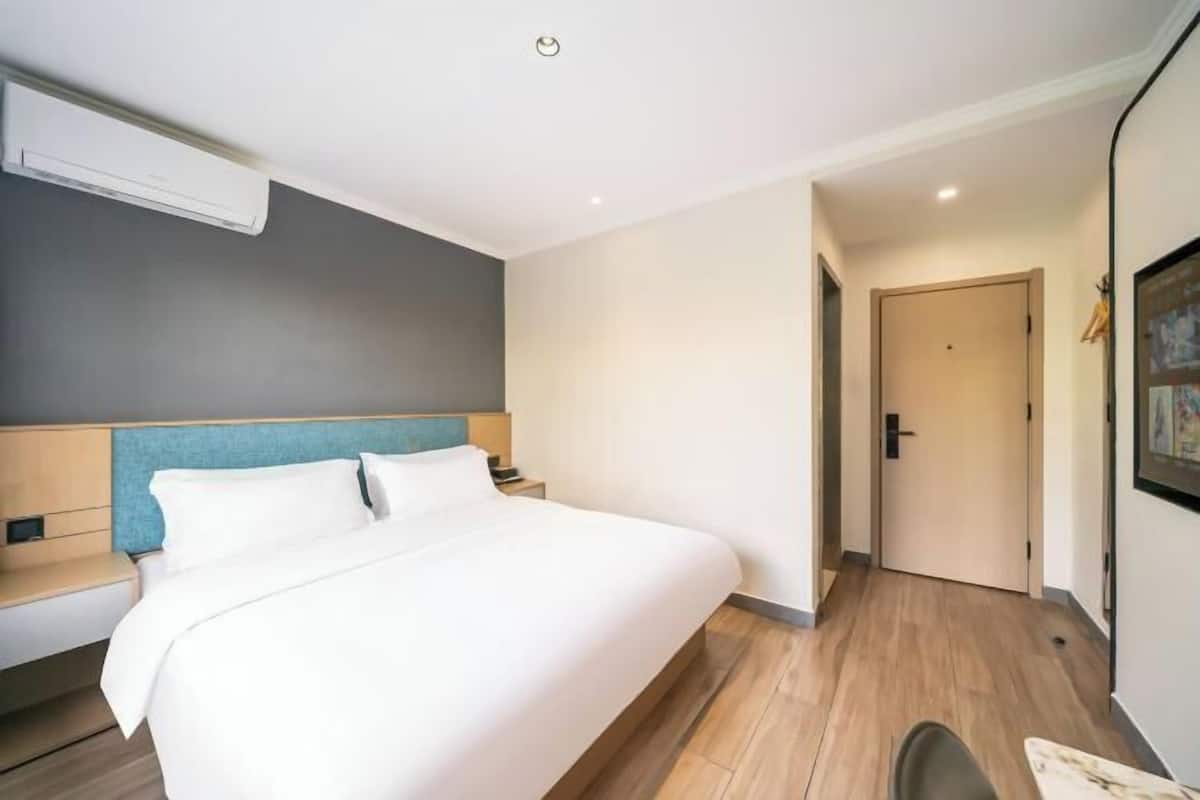 A modern hotel room with a large bed, a bedside table, a nightstand, a wall-mounted air conditioner, and a door leading to another room.