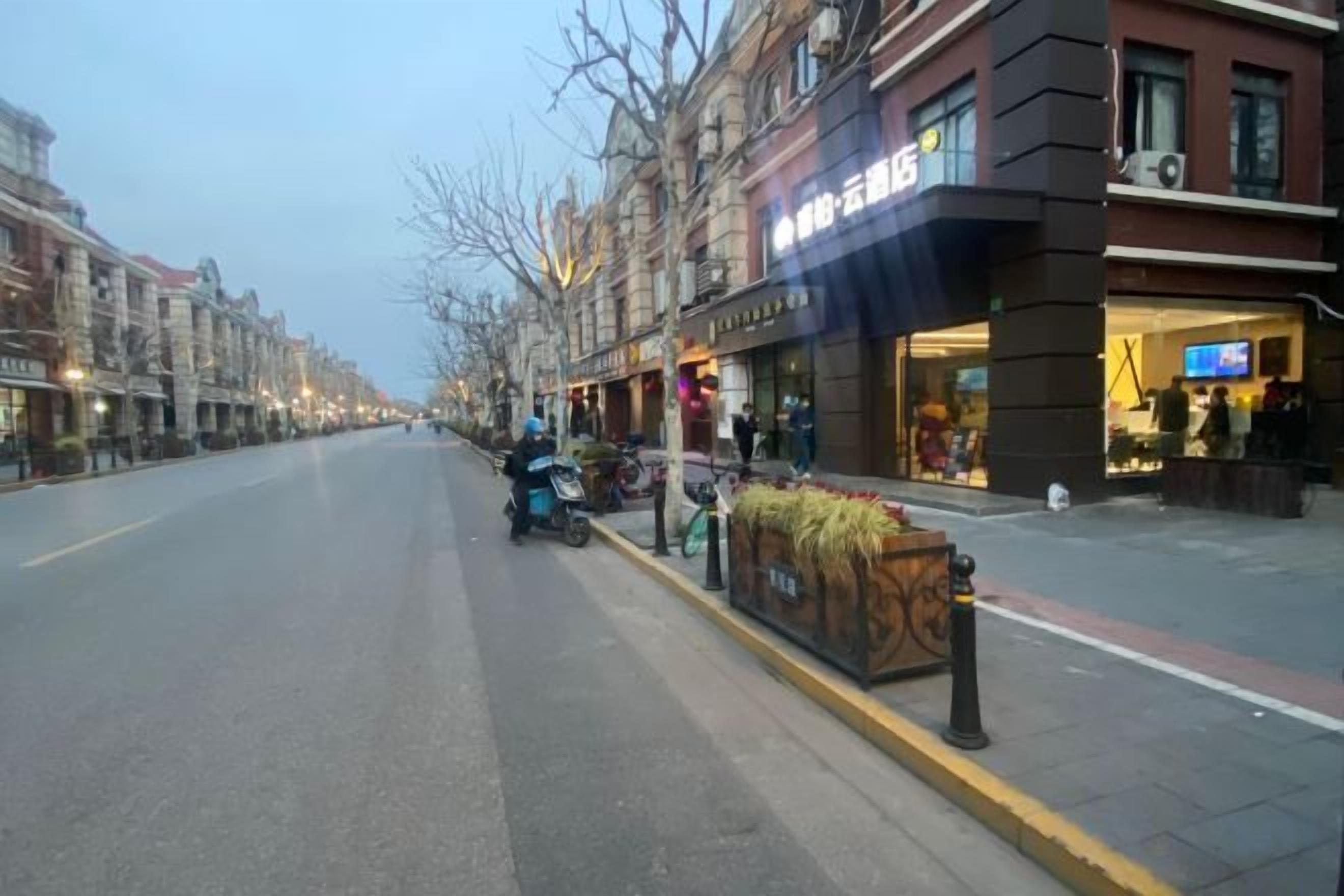 Una calle flanqueada por edificios, una scooter estacionada en la acera, y un local comercial con letreros iluminados.