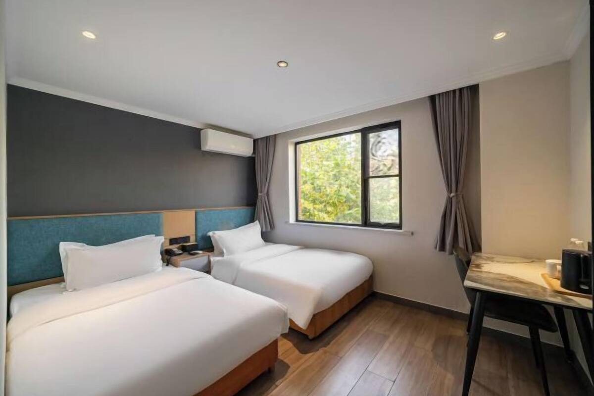 A hotel room with two beds, a desk, a chair, a window with curtains, and an air conditioning unit.