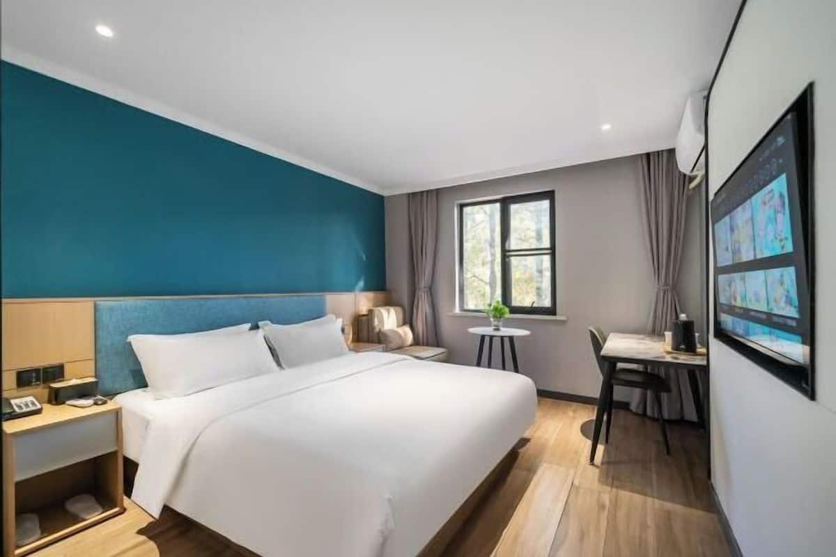 A hotel room with a bed, bedside table, lamp, window, and a desk with a chair.