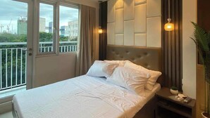 Habitación de hotel con una cama grande, un equipo de aire acondicionado, una mesita de noche, una planta en maceta y vistas a la ciudad a través de la ventana.
