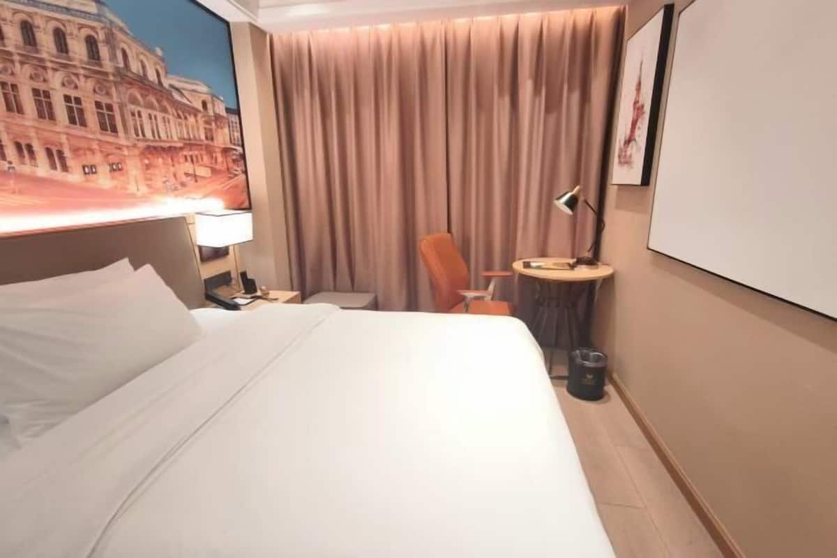 A hotel room with a bed, a desk, a chair, a lamp, a picture frame, and a curtain.