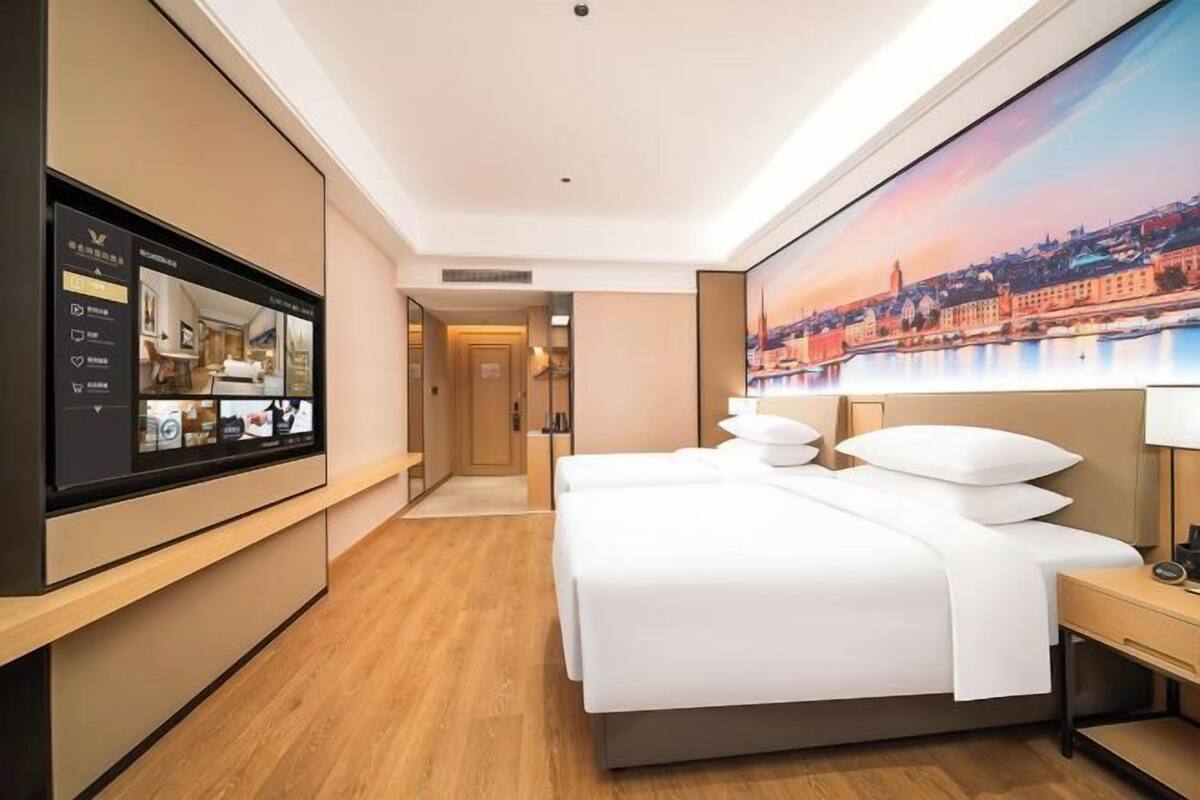 A hotel room with a large bed, a TV on the wall, and a cityscape painting on the wall.