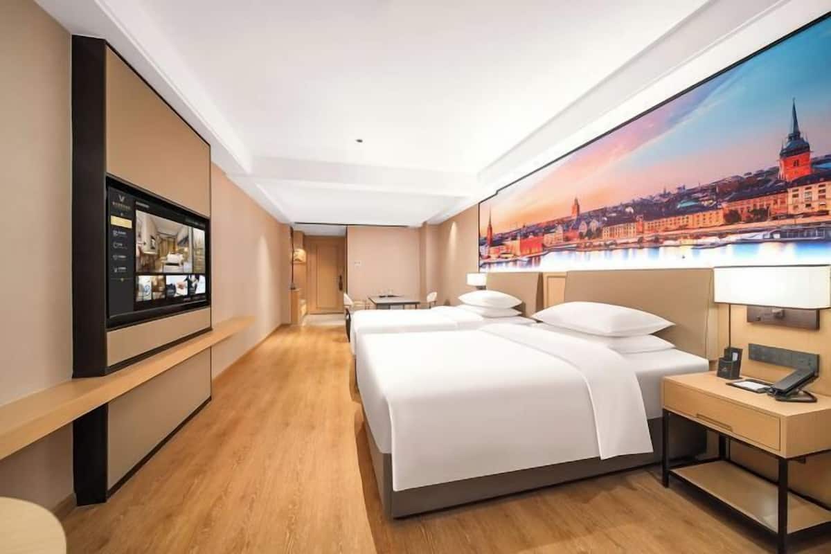 A hotel room with two beds, a flat-screen TV, a painting of a cityscape, and a desk with a lamp.