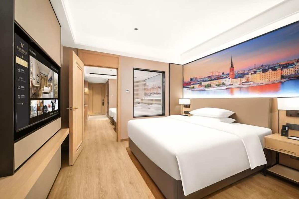 A hotel room with a large bed, a television, a desk, and a cityscape painting on the wall.