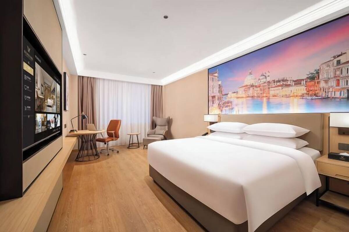 A hotel room with a bed, a desk with a chair, a TV, and a painting of a cityscape.