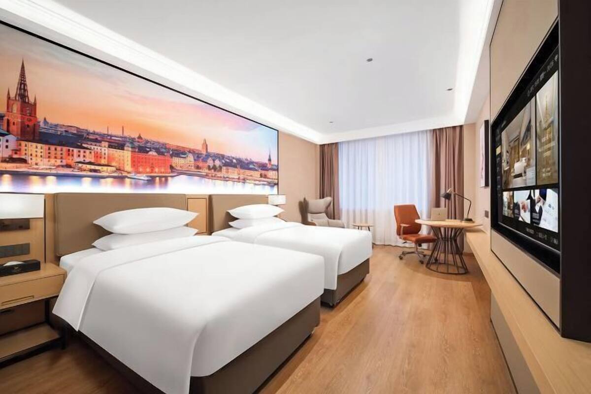 A hotel room with two beds, a desk, a chair, a large window, and a cityscape painting.
