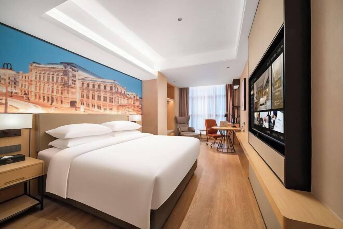 A hotel room with a large bed, a desk, a chair, a TV, and a mural of a building.