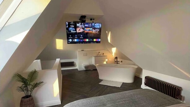 A modern, compact bedroom with a large bed, a TV mounted on the sloped ceiling, a small seating area, and a radiator. - Branksome 4-house Sleeps 8 With Hot Tub & Parking (Worcester)