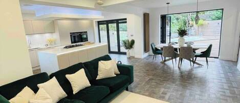 A modern living room with a green sofa, a dining area with a table and chairs, and a kitchen in the background.