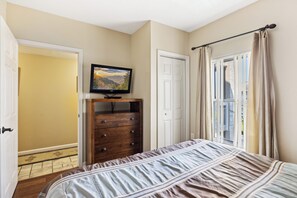 Une chambre à coucher avec un lit, une commode, un téléviseur fixé au mur et une fenêtre avec des rideaux.
