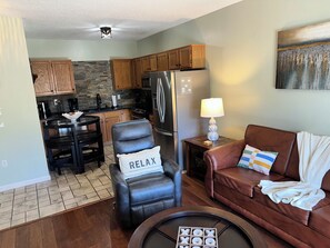 Un espacio habitable compacto con kitchenette, un sillón de cuero con almohada que dice 'RELAX', un sofá de cuero marrón y una mesa de centro con un juego.