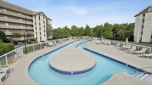 A swimming pool with a curved design, surrounded by lounge chairs and umbrellas, with a multi-story building in the background.