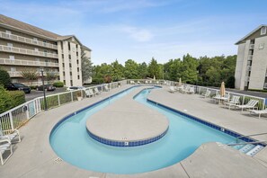 Ein Pool mit geschwungener Form, umgeben von Liegestühlen und Sonnenschirmen, mit einem mehrstöckigen Gebäude im Hintergrund.