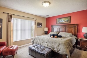 A bedroom with a bed, a nightstand, a lamp, a window with curtains, a red wall, and a painting.