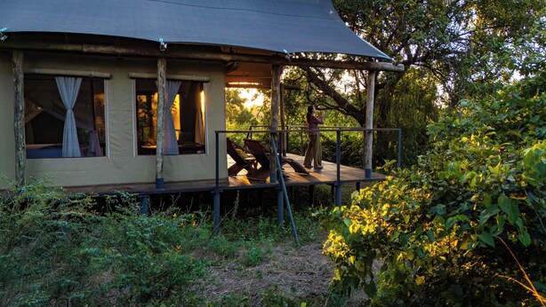A rustic cabin with a covered porch, wooden deck, and a view of the surrounding forest. - Camp Sabali & Safaris (South Luangwa National Park)