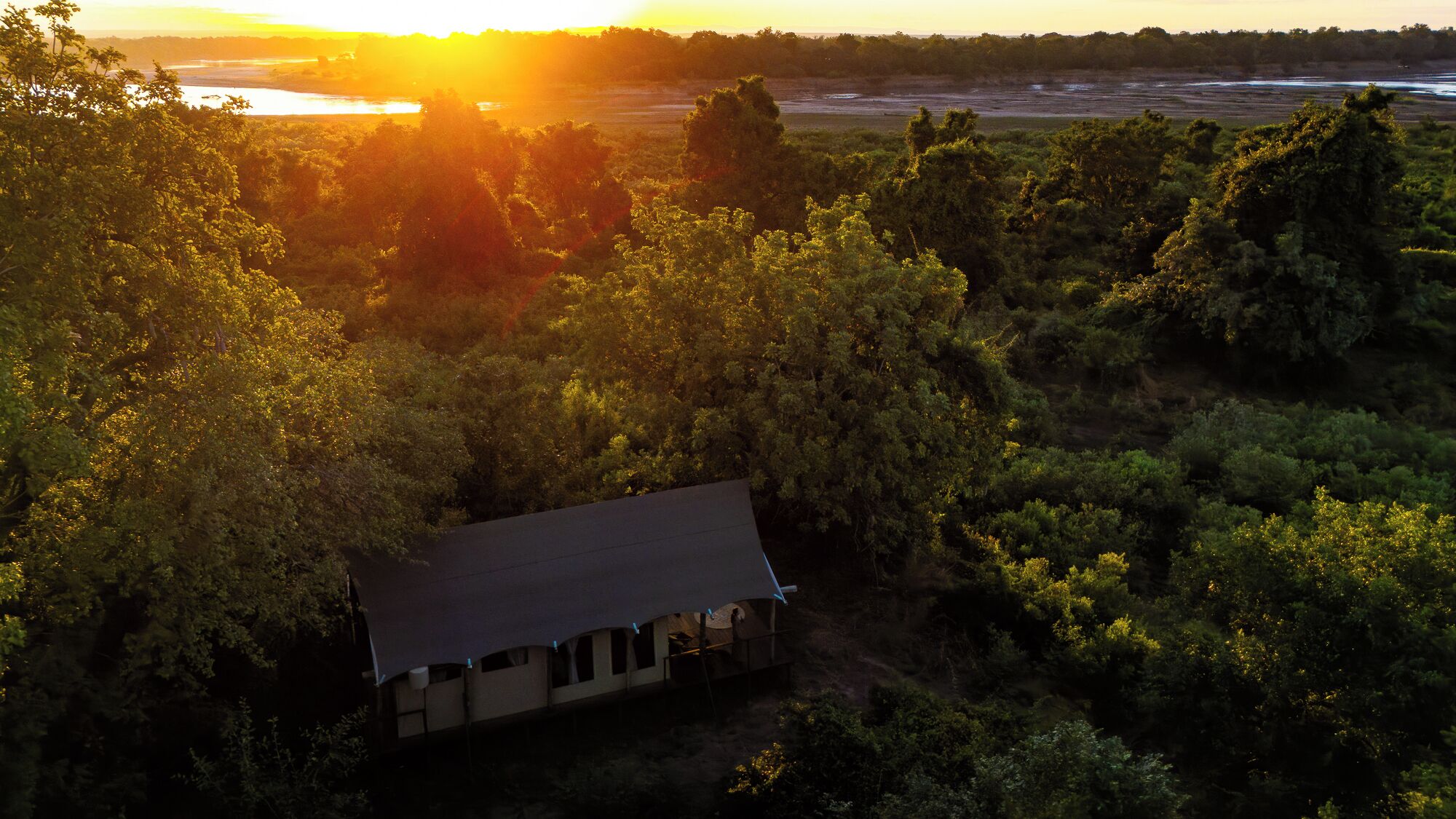 A house surrounded by dense forest with a view of a body of water and a sunset in the background.
