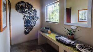 A bathroom with a sink, a decorative wall piece, a window, and a mirror. - Camp Sabali & Safaris (South Luangwa National Park)