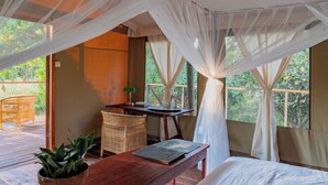 A room with a bed, a wooden table, a wicker chair, a potted plant, and a view of the outdoors through sheer curtains. - Camp Sabali & Safaris (South Luangwa National Park)