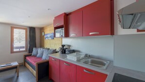Une cuisine compacte avec des meubles rouges, un évier et une cuisinière. On y trouve une petite table à manger et un canapé d’où l’on peut admirer un paysage enneigé par la fenêtre.