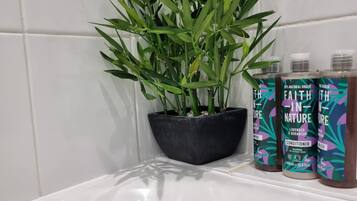 Casa de banho com banheira branca, planta num vaso preto e três frascos de champô e condicionador 'Faith in Nature'.