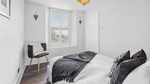 A bedroom with a bed, a chair, a window with blinds, a door, and a wall-mounted light fixture. - Peaceful, Homely, Pet Friendly with Free Parking - Holiday Apartment 13A (Torquay)