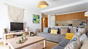 Una sala de estar moderna con sofá, mesa de centro y televisor. Se observa una zona de cocina al fondo.