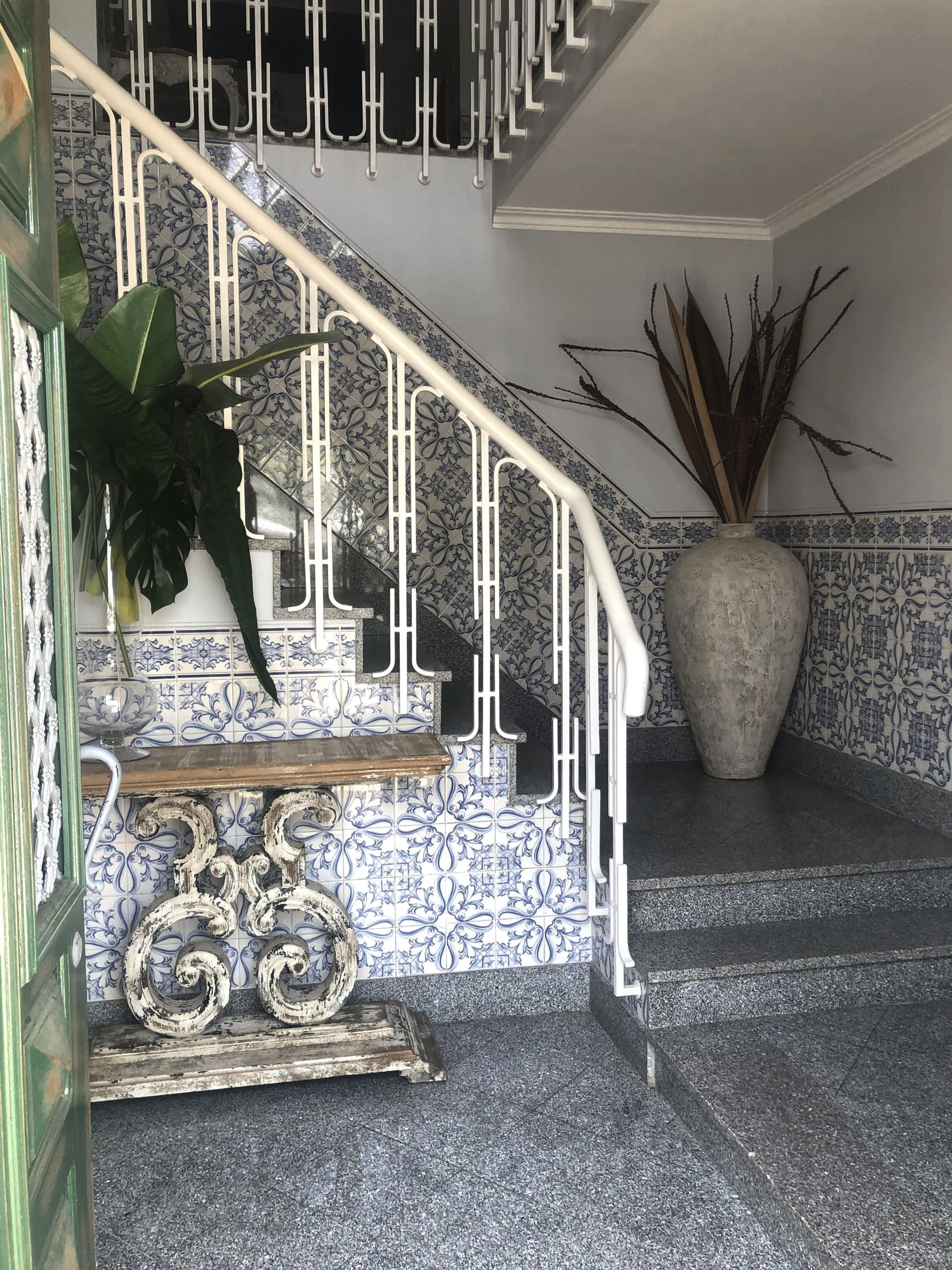 A staircase with a white railing, blue and white patterned tiles, a large potted plant, and a decorative metal piece on the landing.