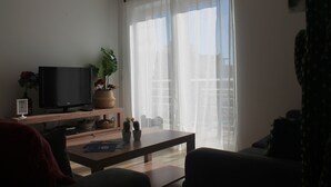 Una sala de estar con un televisor sobre un mueble de madera, una mesa de centro y un sofá. Hay una ventana con cortinas ligeras y vistas a edificios.