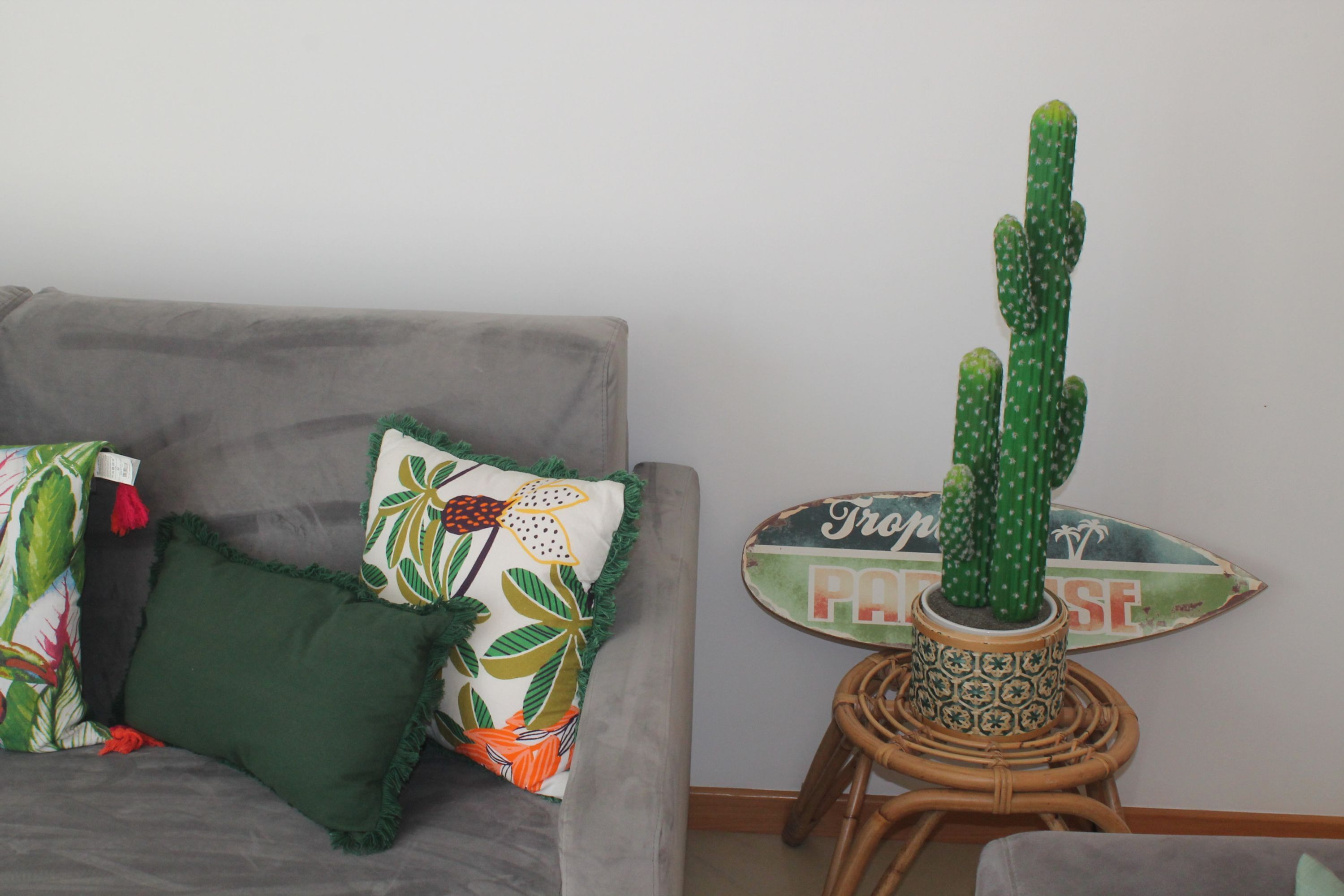 Een grijze bank met groene en oranje kussens, een cactus in een decoratieve pot op een rieten kruk, en een bord met de tekst 'PASTA'.