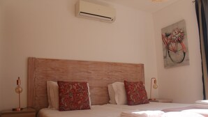 Een net opgemaakt bed met een houten hoofdbord, twee decoratieve kussens, een wandairconditioner, een bedlamp en een schilderij van een fiets.
