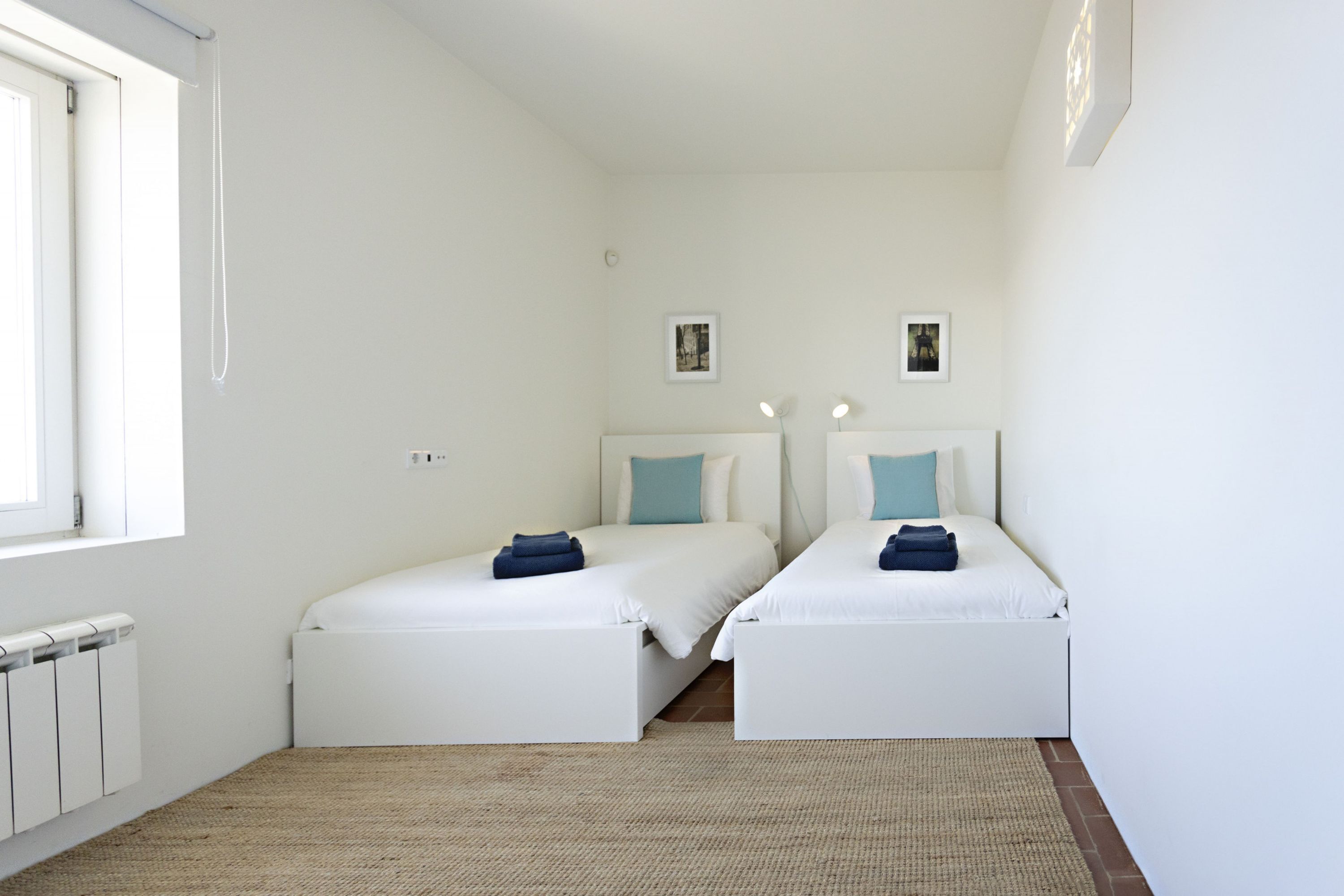 Une chambre avec deux lits simples, un radiateur, une fenêtre et un petit tableau accroché au mur.
