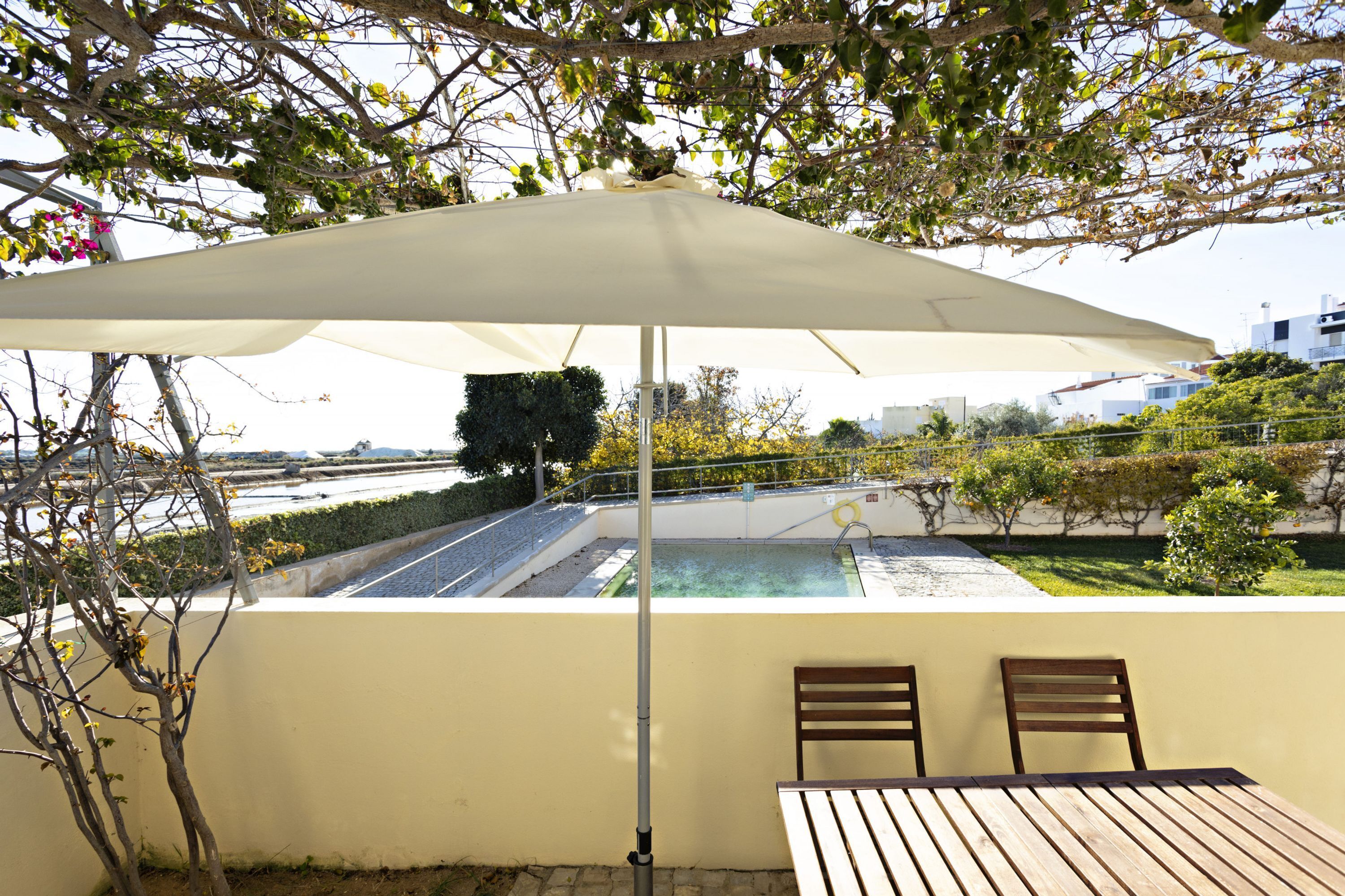 An outdoor seating area with a table and chairs under a large umbrella, overlooking a swimming pool and a landscaped garden.