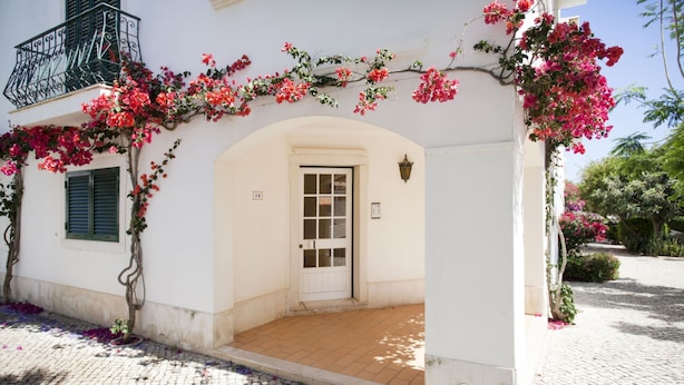 A white building with a balcony, a door, and a small garden area with pink flowers. - Cabanas Village in Tavira (Tavira)