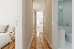 A hallway with a wooden floor leading to a bedroom with a bed and a chair. A bathroom with a toilet and sink is visible to the right.