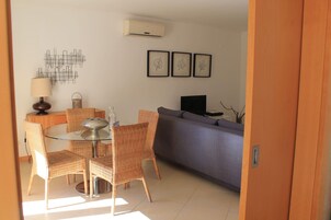 Uma área de refeições com cadeiras de vime e mesa de vidro, um sofá cinzento e arte com folhas emolduradas na parede.