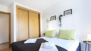 Ett hotellrum med en säng, två kuddar, ett sängbord, en vägghylla med en växt och en dörr.