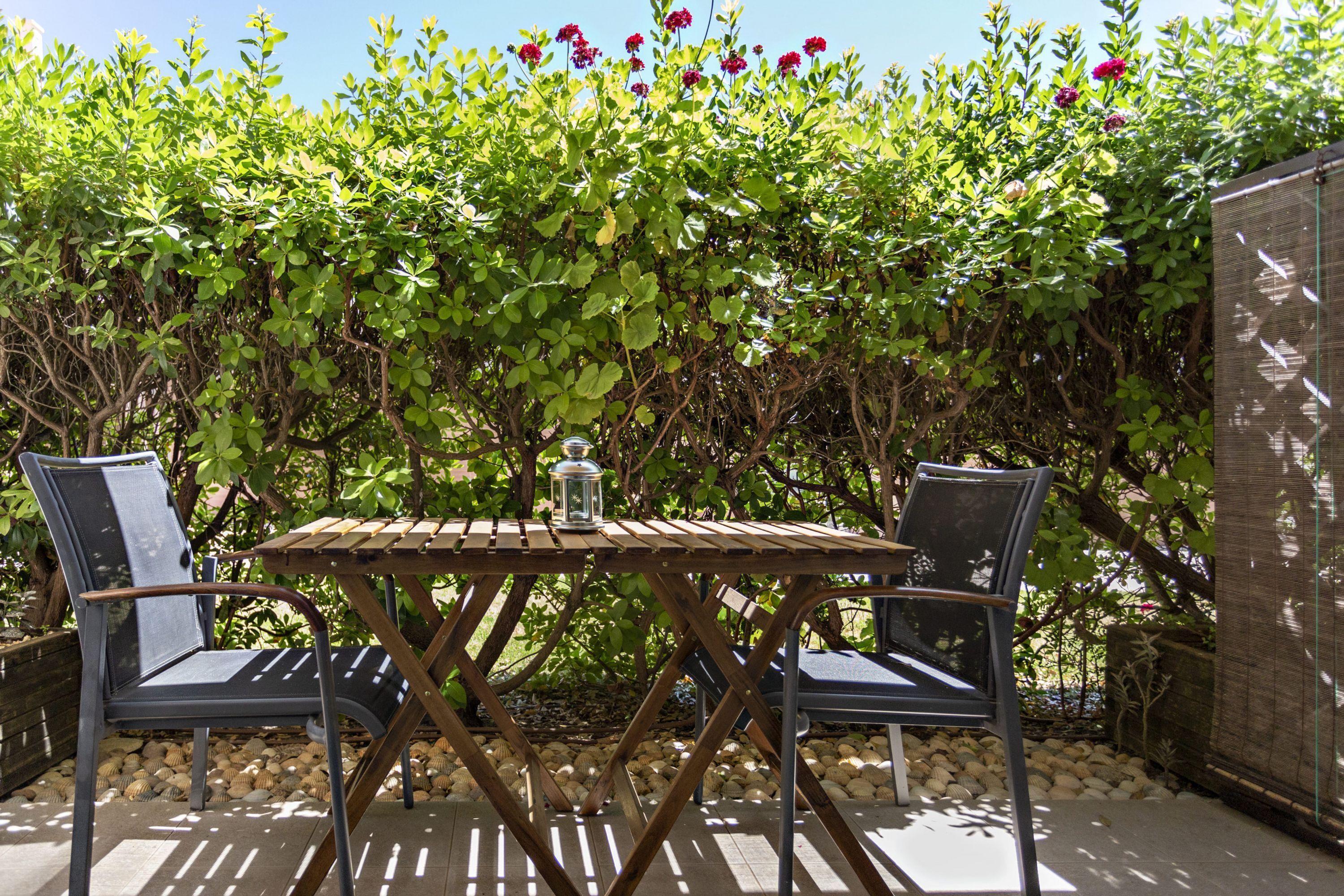 Area esterna con posti a sedere, due sedie e un tavolo, circondata da un giardino con vegetazione verde e fiori rossi.