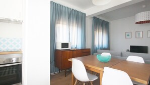 A compact kitchen with a microwave, oven, and wooden cabinet. A dining table with white chairs and a blue bowl is in the center.