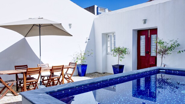 Une piscine aux carreaux bleus, une porte rouge et un bâtiment blanc avec un parasol bleu et des chaises en bois.