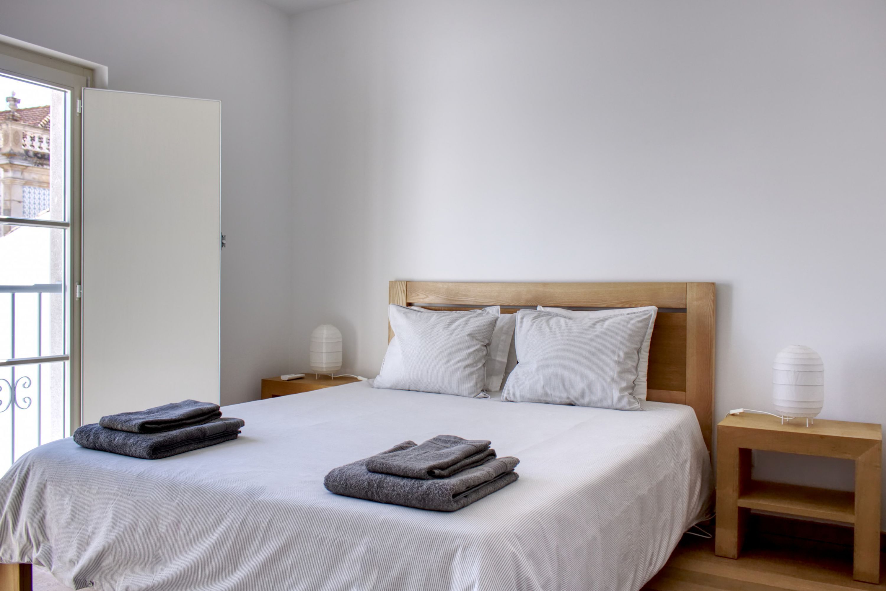 A neatly made bed with a wooden headboard, two bedside tables with lamps, and a view of a building through a window.