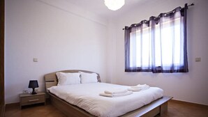 Una cama bien hecha con almohadas y toallas dobladas, una mesita de noche con lámpara y una ventana con cortinas.