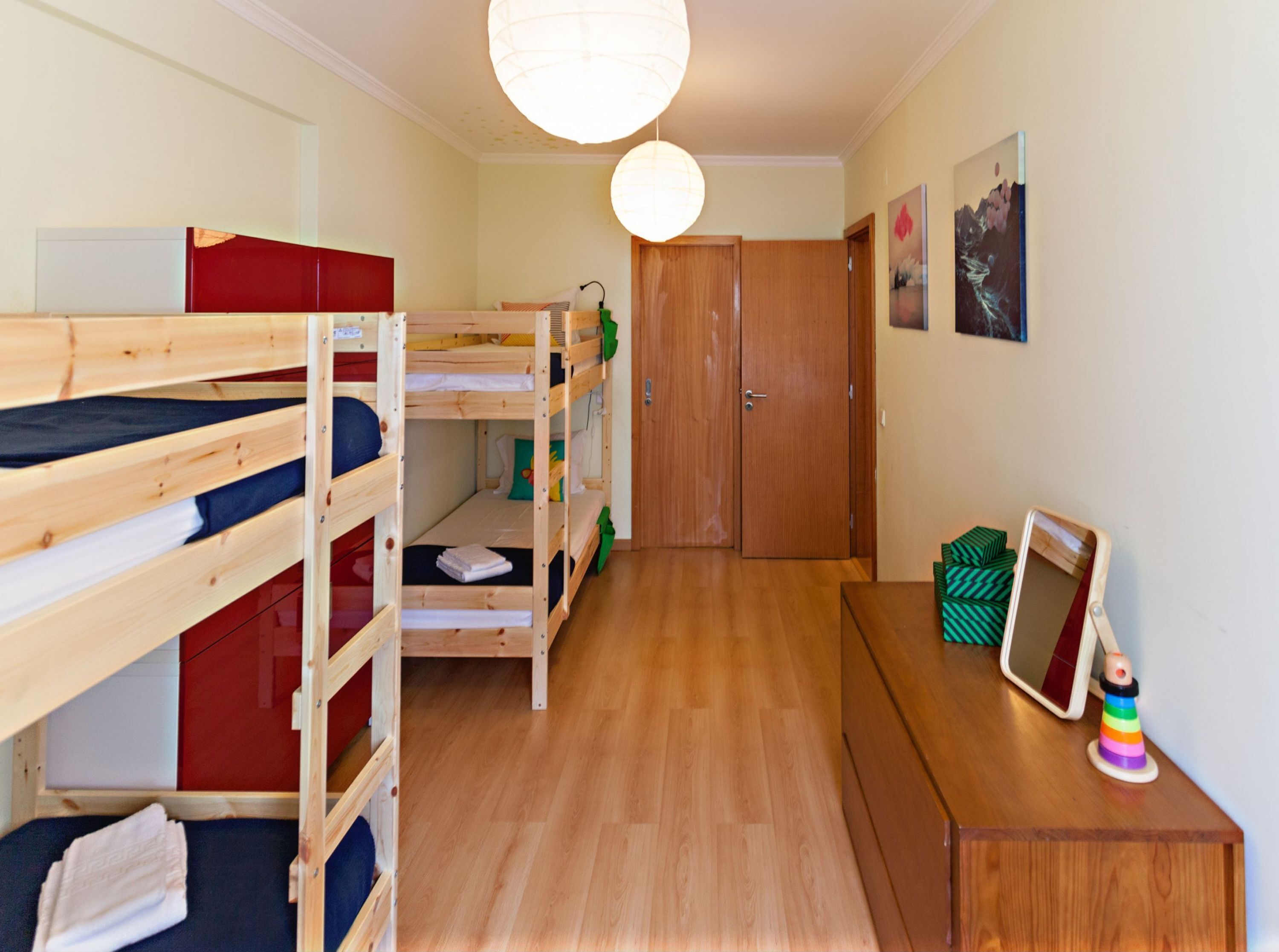 A dormitory room with bunk beds, a wooden wardrobe, a desk with a mirror, and a hanging light fixture.